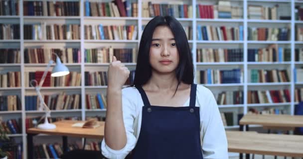 Porträtt av positiv asiatisk tonåring flicka student med svart hår ler mot kameran och visar tummen upp skylt i biblioteket bredvid bokhyllan. — Stockvideo