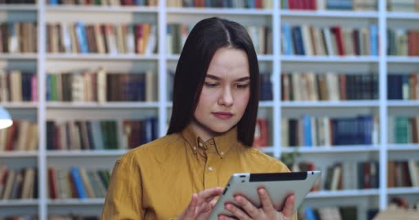 Portret de linda chica en suéter amarillo y pelo largo negro recogiendo datos de su tableta y sonriendo en la biblioteca . — Vídeos de Stock