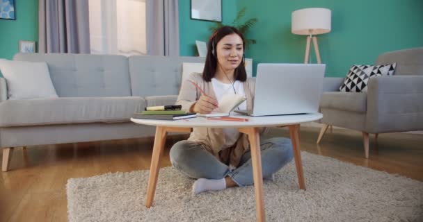 Joven freelancer inspirado trabajando en el ordenador portátil, hablando con los clientes con auriculares. Mujer buscar información y leer noticias en la red social utilizando ordenador portátil en el interior del hogar . — Vídeo de stock