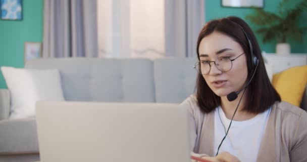 Representante de ventas femenina profesional en auriculares hablando con el cliente, haciendo videollamada, mirando a la computadora portátil, escribiendo notas, hablando por webcam. Vender en línea, trabajar desde casa, telemarketing . — Vídeos de Stock