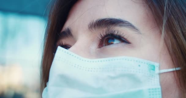 Primo piano di una giovane donna che guarda in camera con gli occhi pieni di speranza e indossa una maschera protettiva sulla strada accanto al centro sanitario. Concetto di salute, vita di sicurezza, coronavirus, protezione dal virus, pandemia — Video Stock