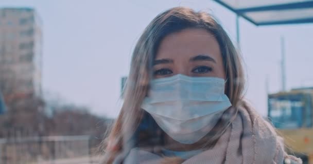 Close up retrato de jovem loira voluntária em uma máscara médica à espera de transporte na parada e olhando para a câmera. Quarentena COVID-19 na Europa. Transmissão do vírus da coronária nos transportes públicos . — Vídeo de Stock