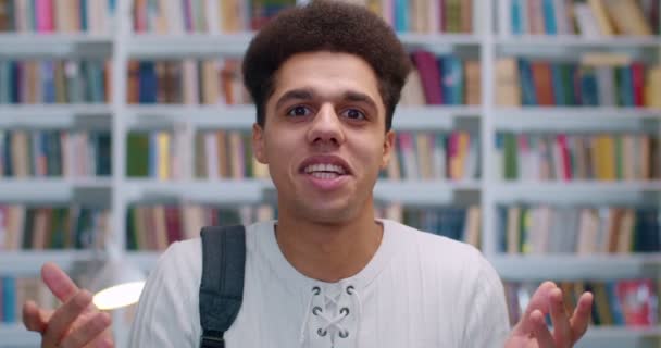 Close-up van Latino jonge knappe man voor de camera staan en vertellen iets emotioneel met de handen gebaren in de bibliotheek. Man student praten met iemand op boekenplanken achter. — Stockvideo