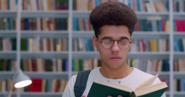 Joven chico guapo latino en gafas leyendo libro de texto en la biblioteca. Primer plano del estudiante masculino con libro en manos estudiando en biblioteca pública con estantes de libros sobre fondo. Concepto de estudio . — Vídeos de Stock