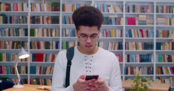 Jeune Latino beau mec dans des lunettes debout et tapant sur smartphone dans la bibliothèque. Joli étudiant masculin avec téléphone dans les mains textos message à bibliotheca avec des étagères de livres derrière . — Video