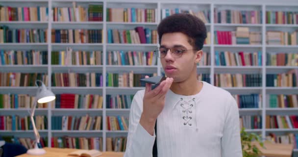 Joven estudiante latino en gafas hablando y grabando mensajes de voz en un smartphone. Tipo guapo enviando grabación de audio de voz o notificación por teléfono en la biblioteca . — Vídeo de stock