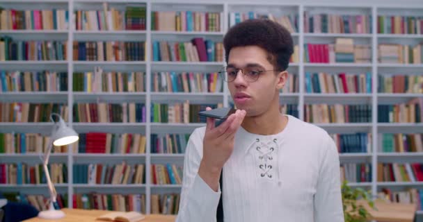 Férfi fiatal latin diák szemüvegben beszél és hangüzenetet rögzít az okostelefonon. Jóképű srác küld audio felvétel beszéd vagy értesítés telefonon keresztül a könyvtárban.