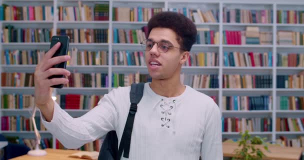 Beau jeune homme Laino dans des lunettes debout dans la bibliothèque et ayant vidéochat sur smartphone. Étudiant masculin parlant via webcam au téléphone avec des étagères de livres sur fond . — Video