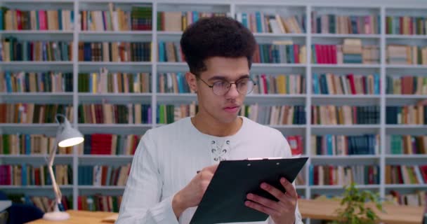 Joven chico guapo latino con gafas de pie en la biblioteca con documentos en la carpeta, anotando y escribiendo algo. Estudiante masculino que estudia en bibliotheca mientras revisa algunos documentos . — Vídeos de Stock