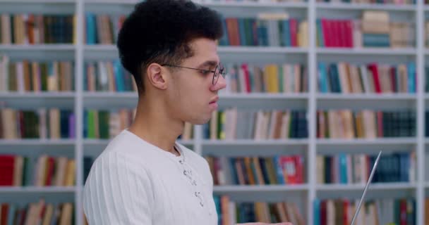 Boční pohled na studenta v brýlích, který drží notebook a píše na klávesnici v knihovně. Detailní záběr na latino chlap otáčení tváří v tvář fotoaparátu a usmívání v bibliotheca při studiu. — Stock video