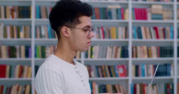 Zijaanzicht op mannelijke student in glazen met laptop computer en typen op toetsenbord in de bibliotheek kamer. close-up van latino guy draaien gezicht naar camera en glimlachen in bibliotheca terwijl studeren. — Stockvideo