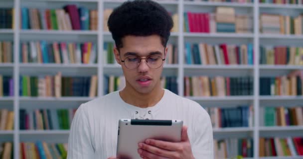 Latino jovem estudante bonito do sexo masculino em óculos de pé na biblioteca e segurando dispositivo tablet na sala da biblioteca. Um tipo a ver algo no computador tablet em Bibliotheca. Estudo do conceito . — Vídeo de Stock