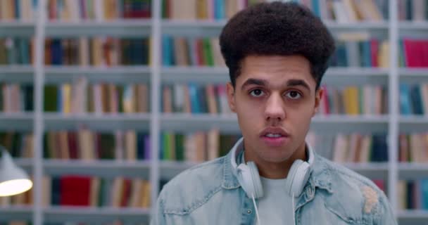 Zbliżenie młodego latynoskiego studenta ze słuchawkami stojącymi w bibliotece i patrzącymi prosto w kamerę. Portret stylowego faceta w bibliotece z półkami na książki na tle. — Wideo stockowe