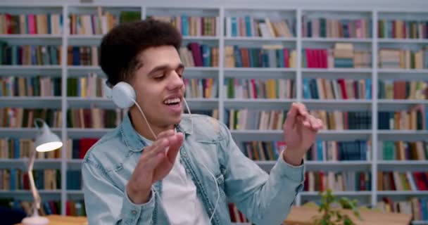 Radostný mladý Hispánec v bílých velkých sluchátkách poslouchající hudbu a vesele tančící před kamerou v knihovnickém prostoru. Muž student odpočinku a bavit se na veřejnosti bibliotheca. — Stock video