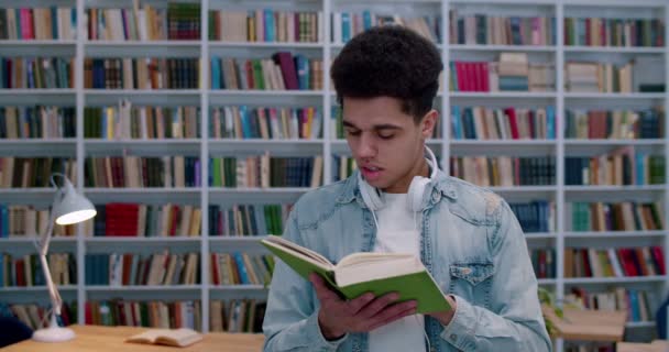 Fiatal latin diák tankönyvet olvas a könyvtárban és tanul. Egy jóképű fickó portréja fejhallgatóval, aki bezárja a könyvet, és a bibliotheca térben mosolyog a kamerába..