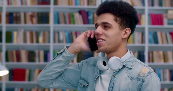 Primer plano de un joven estudiante latino de pie en el espacio público de la biblioteca y hablando por teléfono móvil como si descubriera algo. Un tipo guapo hablando por celular en Bibliotheca. Conversación telefónica . — Vídeos de Stock