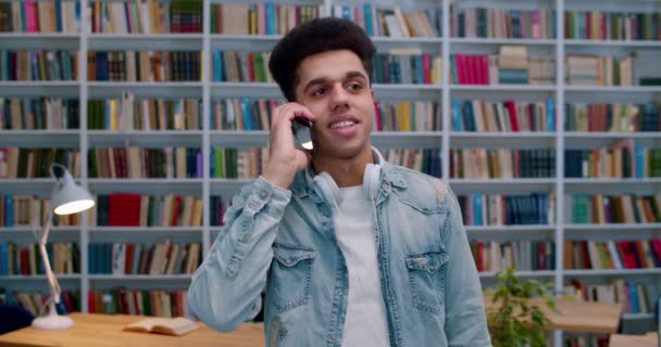 Jonge Latino mannelijke student in de bibliotheek kamer en praten op mobiele telefoon en glimlachen. Knappe vent die op een mobieltje spreekt in bibliotheca. Telefoongesprek. Studie concept. — Stockvideo