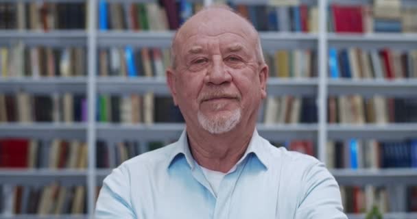 Ritratto di vecchio uomo calvo caucasico in piedi in libreria e guardando la macchina fotografica. Primo piano del professore o operaio di biblioteca. Libri scaffali su sfondo. Insegnante senior . — Video Stock