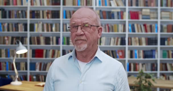 Retrato de hombre mayor calvo caucásico en gafas girando cara a cámara y sonriendo. Primer plano del profesor masculino o profesor posando en bibliotheca. Libros estantes detrás. Senior sonrió trabajador de la biblioteca . — Vídeos de Stock