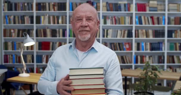 Portrait shot of young handsome bald Caucasian man with stack of textbooks standing in libary and looking at camera. Male senior professor or worker in bibliotheca with books shelves on background. — Stock Video
