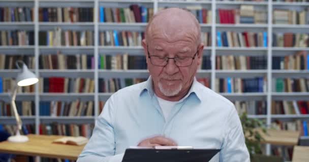 Biały łysy mężczyzna w okularach stojący w bibliotece z dokumentem w folderze, notujący i piszący coś. Mężczyzna bibliotheca pracownik lub starszy nauczyciel sprawdzanie niektórych dokumentów. Profesor pracuje. — Wideo stockowe