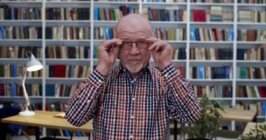 Kütüphane odasında duran, gözlük takan ve kameraya bakan beyaz, kel, yaşlı bir adamın portresi. Erkek öğretmen hafifçe gülümsüyor. Kitaplar rafların arkasında. Bibliyotheca Profesörü