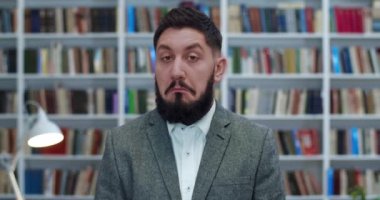 Kütüphanede kameranın önünde şaşırmış yüzüyle duran ceketli, yakışıklı, beyaz adamı yakından çek. Erkek, bir şeyi onaylar gibi başını sallıyor. Konsept çalış.