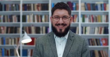Yakışıklı bir adamın libary 'de dikilip kameraya gülen portresi. Bibliyotheca 'daki mutlu erkek yüzüne yakın dur. Kapalı alanda gülümseyen, arka planda kitap rafları olan genç beyaz bir adam.