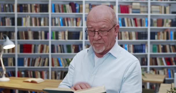 Vecchio uomo calvo caucasico in bicchieri leggere libro in biblioteca. Primo piano del professore di sesso maschile con libro di testo in mano che studia in biblioteca pubblica. Concetto di studio. Nonno sfogliando le pagine . — Video Stock