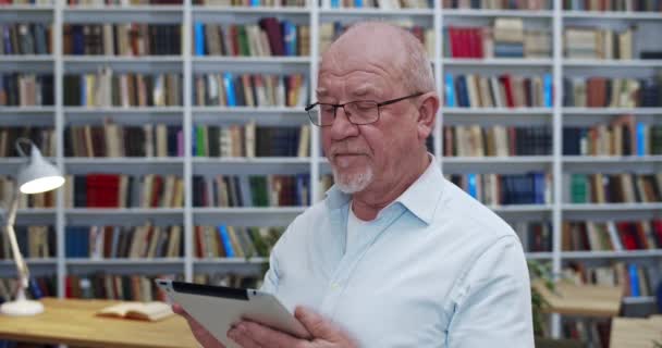 Caucasian old male teacher in glasses standing in library and holding tablet device. Portrait of senior wise man watching something on tablet computer and then smiling to camera in bibliotheca. — Stock Video