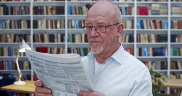 Ritratto di vecchio uomo calvo caucasico in occhiali che legge il giornale in biblioteca e guarda alla macchina fotografica. Primo piano del professore di sesso maschile con gazzetta rivista di ricerca informazioni in bibliotheca . — Video Stock