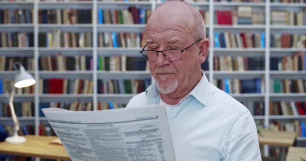 Старий кавказький лисий чоловік в окулярах читає газету в бібліотеці і посміхається. Зблизька професор чоловічої статі з газетним журналом шукає інформацію в бібліотеці. Дід розслідує новини. — стокове відео
