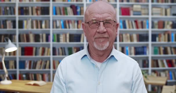 Retrato do velho homem careca caucasiano em óculos olhando para a câmera com um leve sorriso na biblioteca. Close up de professor ou trabalhador do sexo masculino de bibliotheca. Prateleiras de livros no fundo. Professor sénior . — Vídeo de Stock