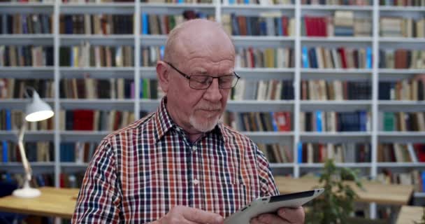 Vieux professeur masculin caucasien dans des lunettes debout dans la bibliothèque et tenant tablette dispositif. Sage aîné regardant quelque chose sur tablette et tapant ou défilant à l'écran dans bibliotheca . — Video