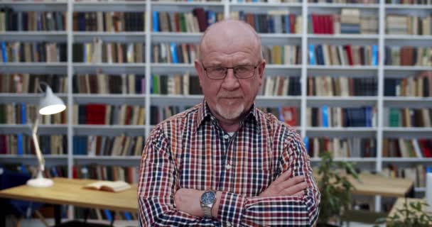 Portrait shot of Caucasian old bald man in glasses and motley shirt with crossed hands standing in library room and looking straight at camera. Books shelves on background. Professor in bibliotheca. — Stock Video