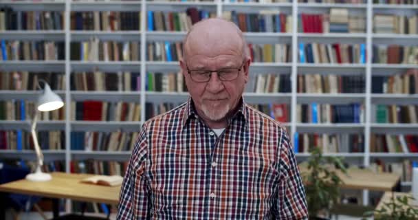 Retrato de hombre calvo viejo caucásico en gafas y camisa a cuadros de pie en la sala de la biblioteca y mirando hacia abajo. Profesor varón levantando los ojos a la cámara. Libros estantes en el fondo. Profesor en bibliotheca . — Vídeo de stock