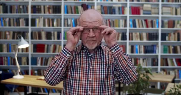 Retrato del viejo calvo caucásico con camisa abigarrada de pie en la sala de la biblioteca, tomando gafas y mirando a la cámara. Profesor senior masculino sonriendo levemente. Libros estantes detrás. Profesor en bibliotheca — Vídeos de Stock