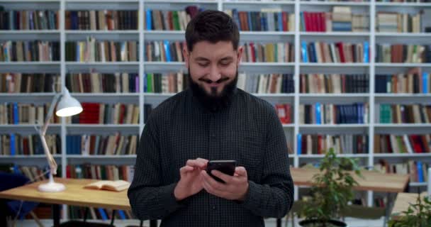 Ung vit stilig man med skägg stående och knacka på smartphone i biblioteket. Snygg manlig student eller forskare med telefon i händerna SMS i bibliotheca med många böcker. — Stockvideo