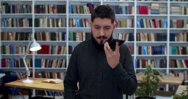 Man unga kaukasiska student eller vetenskaplig forskare talar och spela in röstmeddelanden på smartphone. Snygg man med skägg skicka ljudinspelning av tal eller avisering via telefon i biblioteket. — Stockvideo