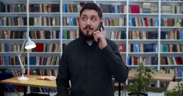 Young Caucasian male student standing in library room and talking on mobile phone and smiling. Handsome man speaking on cellphone in bibliotheca. Telephone conversation. Guy in book store. — Stock Video