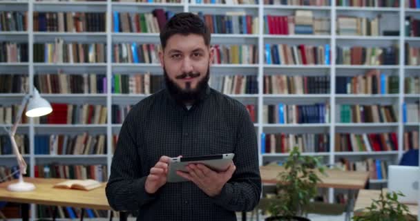 Giovane uomo d'affari caucasico bello in piedi in biblioteca e tenendo dispositivo tablet, utente online. Un uomo che guarda qualcosa al computer e poi guarda la telecamera. Ritratto di ricercatore in biblioteca . — Video Stock