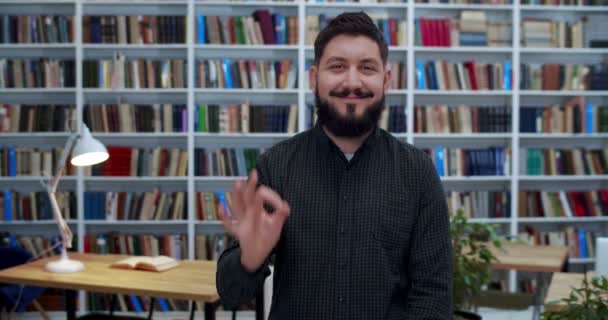 Porträt eines kaukasischen jungen Mannes mit Bart, der fröhlich lächelt und seine Geste mit der Hand im Bibliotheksraum zeigt. Glückliche männliche Schüler oder Lehrer posieren vor der Kamera und freuen sich nach den Prüfungen. Studienerfolg — Stockvideo