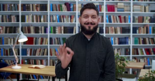 Retrato de un joven caucásico con barba sonriendo alegremente y mostrando un gesto OK con la mano en la sala de la biblioteca. Feliz estudiante masculino o profesor posando ante la cámara y es alegre después de los exámenes. Éxito en el estudio — Vídeo de stock