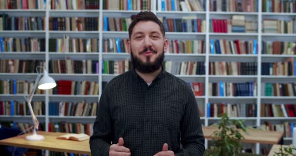 Jeune homme caucasien avec la barbe debout devant la caméra et raconter une histoire émotionnellement avec des gestes de mains à la bibliothèque. Portrait de l'enseignant masculin expliquant le concept. Blogueur vidéo ou coach . — Video