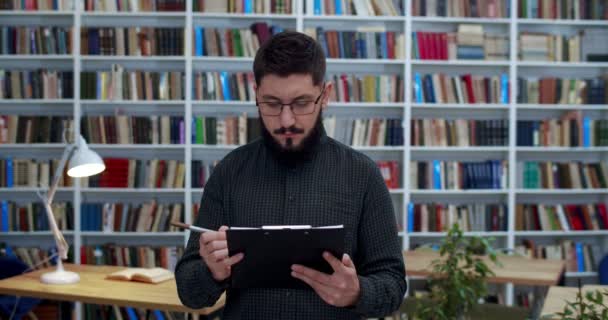 Homem bonito jovem caucasiano de óculos de pé na biblioteca com documentos na pasta, observando e escrevendo algo. Estudante ou pesquisador do sexo masculino que estuda em bibliotheca enquanto verifica alguns artigos . — Vídeo de Stock