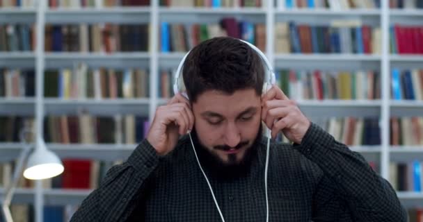 Junger gutaussehender Mann setzt sich Kopfhörer auf und hört im Bibliotheksraum schöne Musik. Nahaufnahme eines kaukasischen männlichen Studenten, der Gesang genießt, während er in der Bibliotheca ruht und viele Bücher hinter sich hat. — Stockvideo