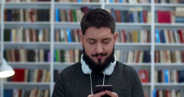 Young Cucasian bărbat bibliotecă lucrător sau cu căști atingând și mesaje text pe telefonul mobil. Omul tastează și defilează pe smartphone în bibliotheca. Magazin de carte costomer chat cu mesaje . — Videoclip de stoc