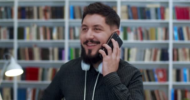 年轻的白人男生，戴着耳机，一边用手机聊天，一边在图书馆里笑。在书目里用手机说话的英俊而快乐的男人。电话交谈。书店的人. — 图库视频影像