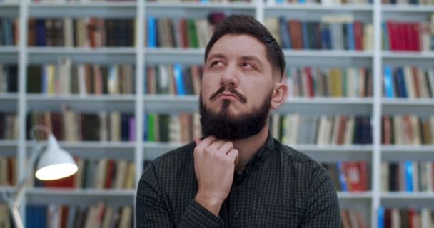 Primo piano di un bell'uomo caucasico in piedi in libreria con la faccia pensierosa. Ritratto di un serio studente o ricercatore di sesso maschile in biblioteca che considera qualcosa e sorride. Libri scaffali dietro . — Video Stock