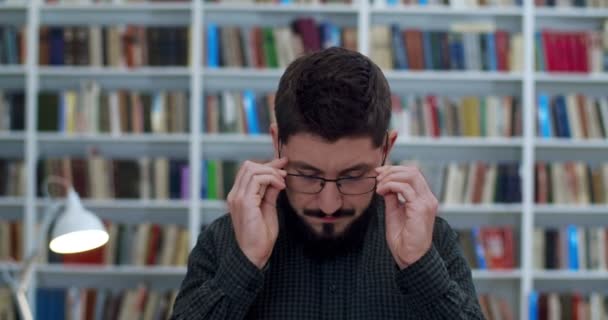 Kütüphanede sakallı, gözlük takan ve kameraya bakan beyaz bir gencin portresi. Erkek öğretmen hafifçe gülümsüyor. Kitaplar rafların arkasında. Bibliyotheca Profesörü. — Stok video